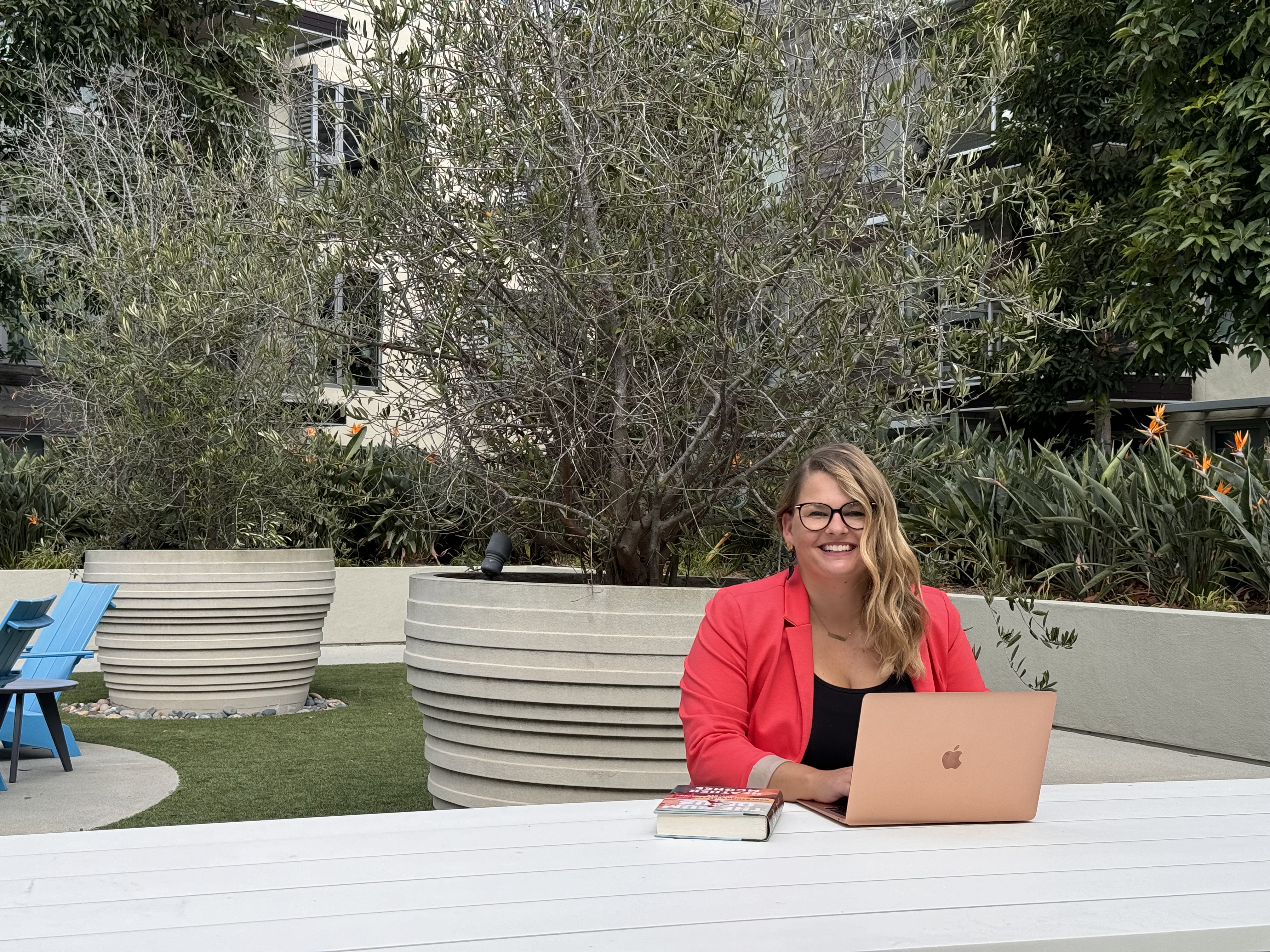 Photo of Lynn Marie Debilzen, founder of The Collective Shift Co., smiling and seated at a white picnic table outside with a laptop