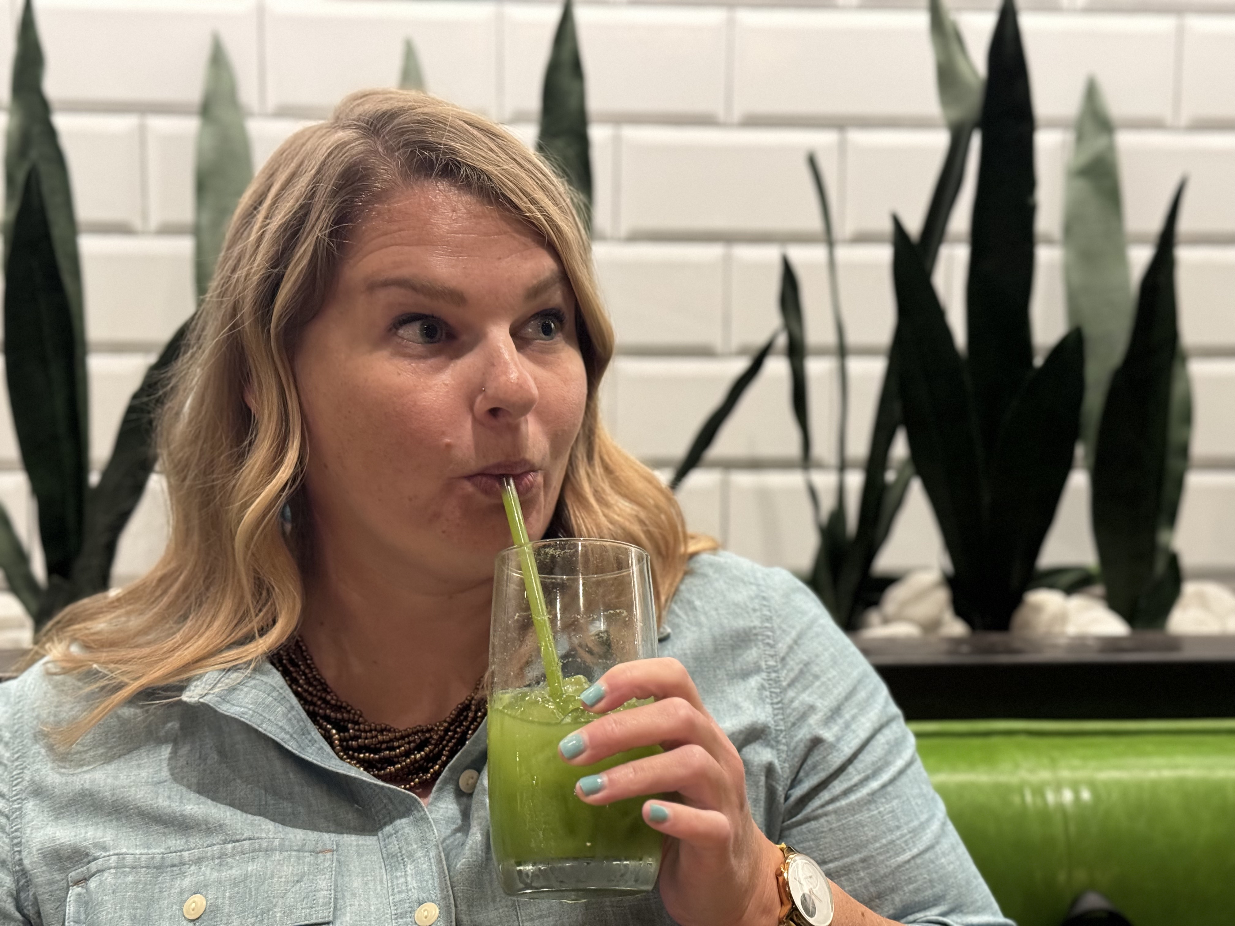 Photo of Lynn Marie Debilzen, founder of The Collective Shift Co., drinking a green juice in front of green plants